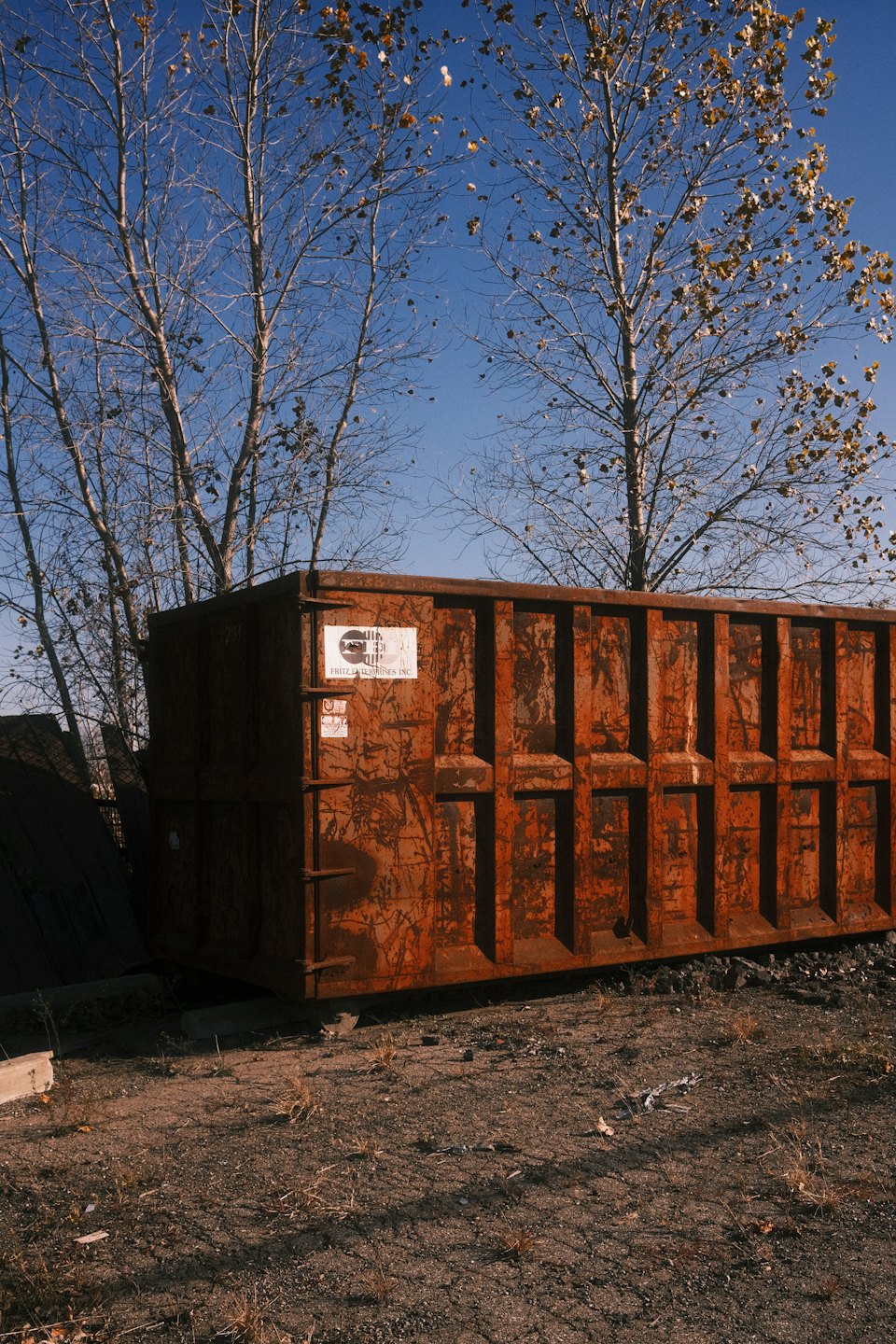 Rusty industrial dumpster sitting on a gravel lot near trees.