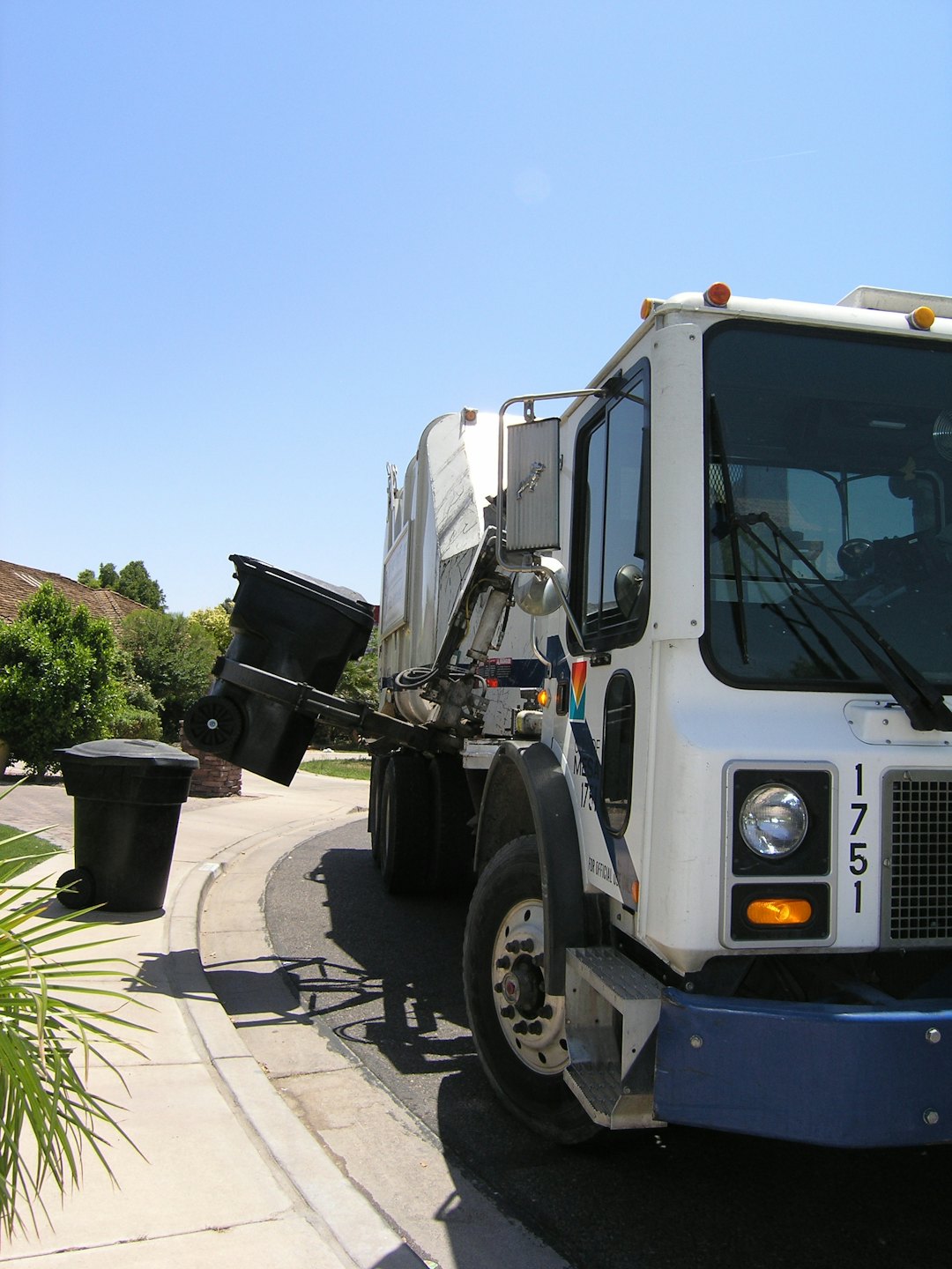 Garbage truck lifts trash bin on a sunny street.