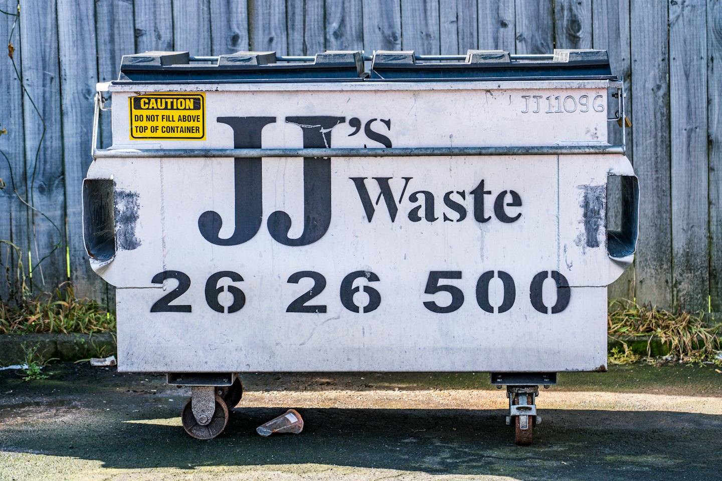 JJ Waste commercial dumpster standing in front of a wooden fence.