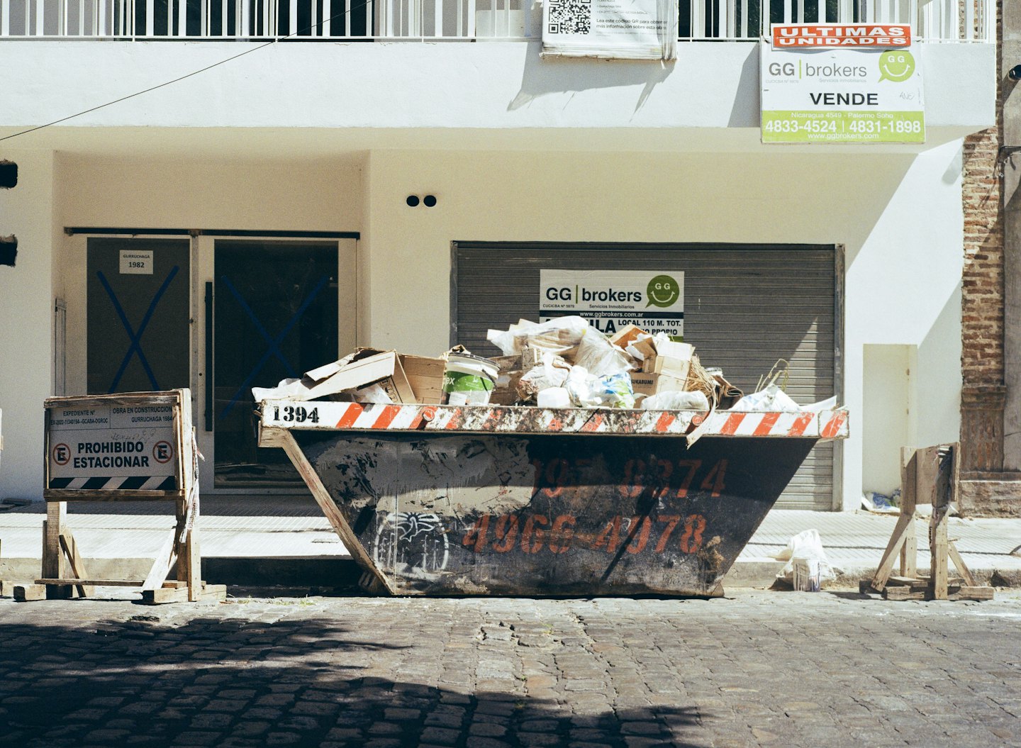 A construction dumpster sits on a cobblestone street by a building.