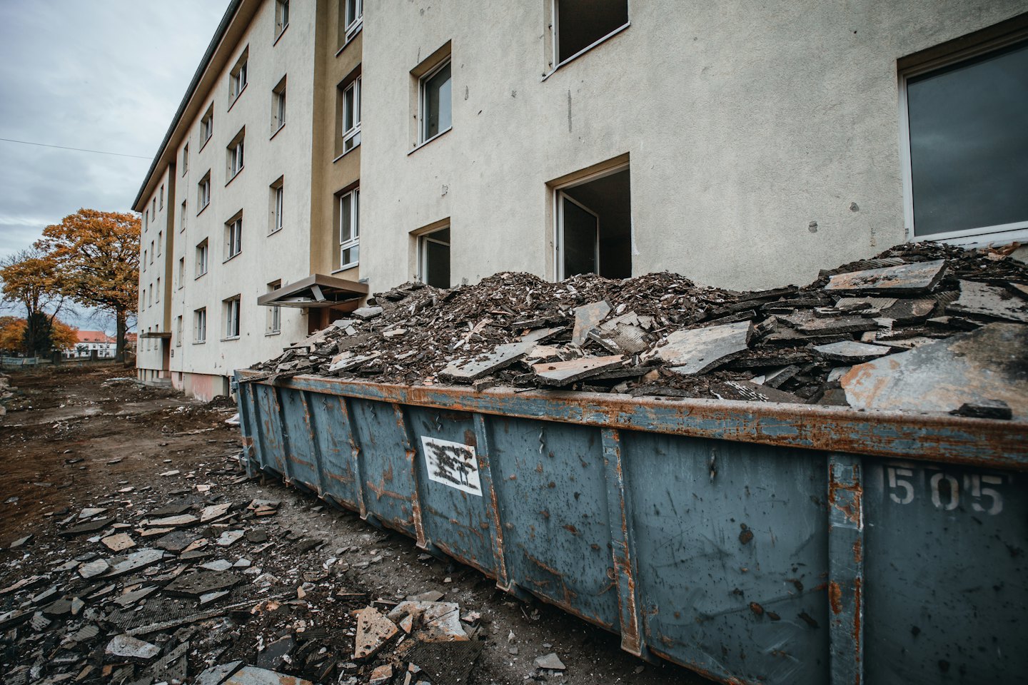 Overflowing blue dumpster at concrete building renovation site.